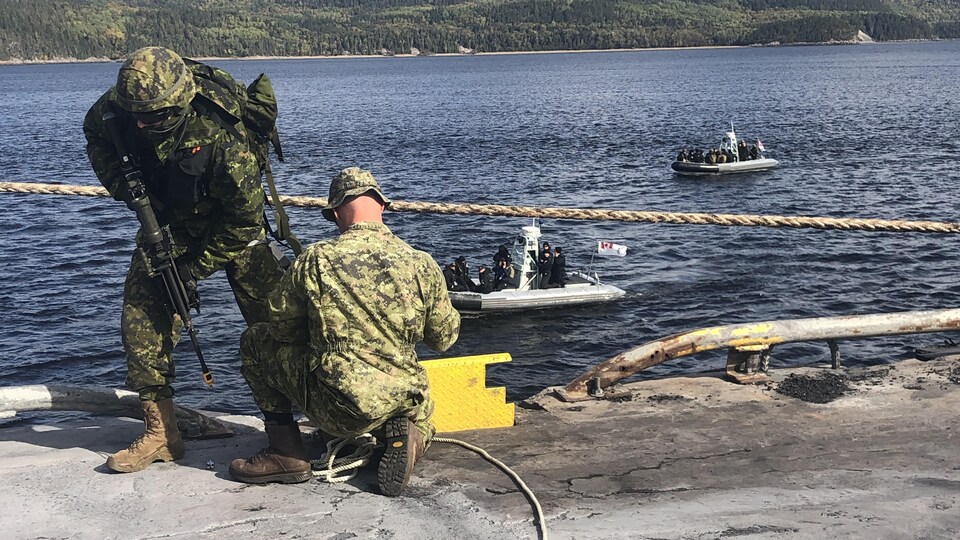 Un militaire en avant plan avec une arme. Un bateau sur la rivière Saguenay à l'arrière-plan.