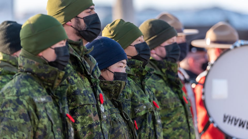 Des militaires en uniforme, en rang, chaudement habillés, la tête couverte d'une tuque et un masque en tissu sur le visage.