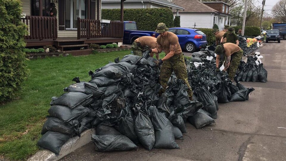 Des militaires ramassent des sacs de sable devant une maison.