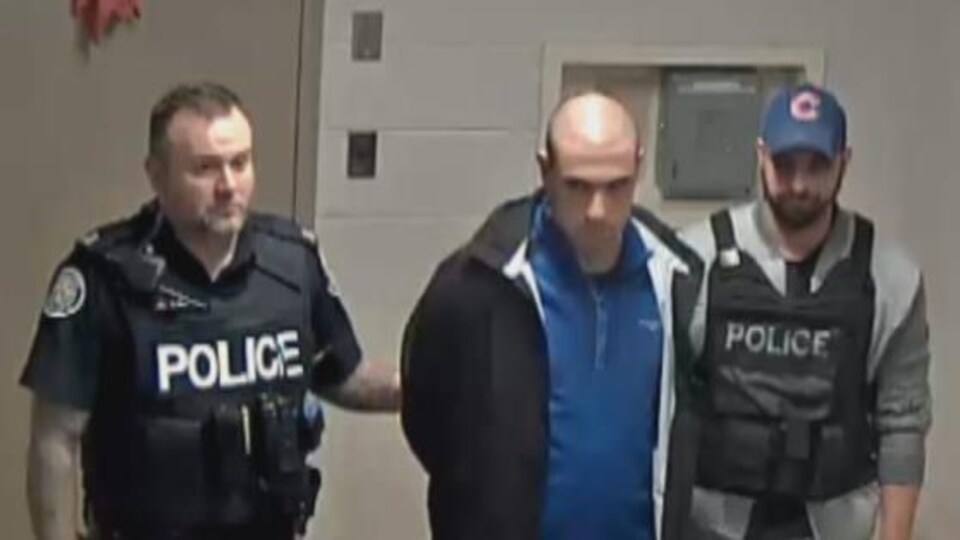 Un homme menotté et escorté par deux policiers.