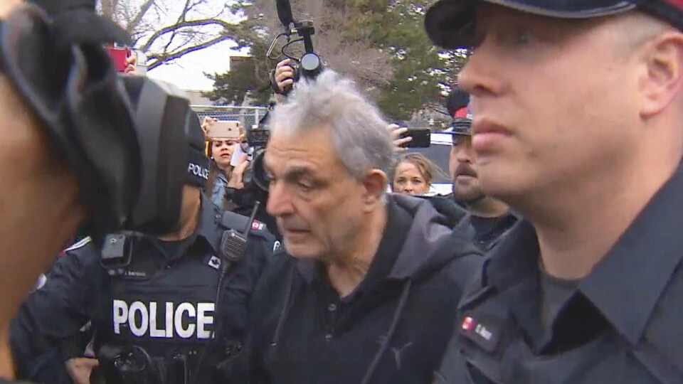 Un vieil homme entouré par des policiers pour le protéger des médias.