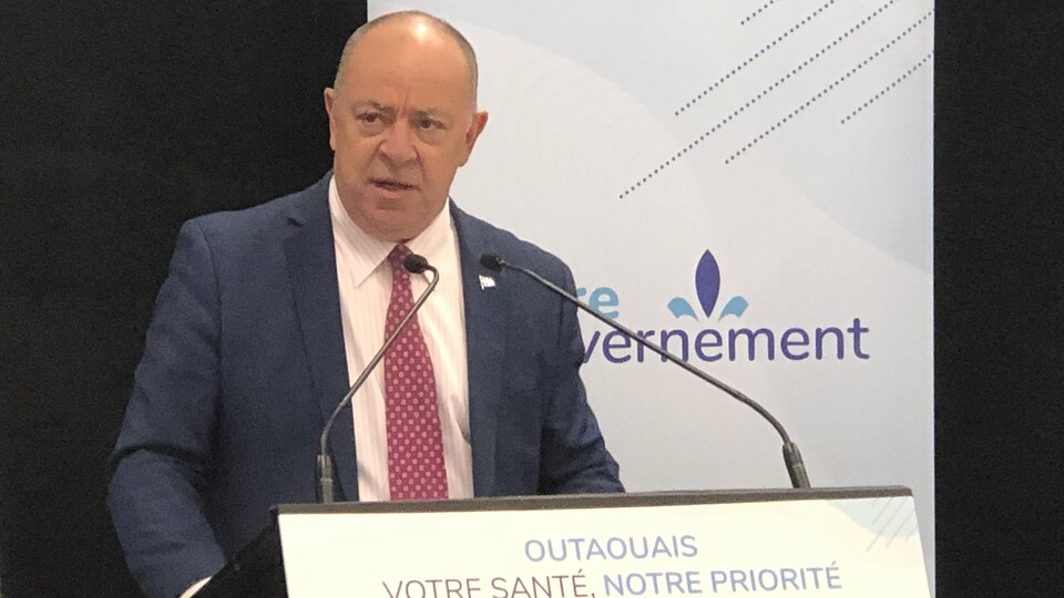 L'homme à la cravate est devant un lutrin sur lequel est ajusté une affiche sur laquelle il est écrit «Outaouais - votre santé, notre priorité».