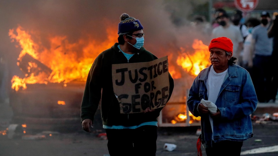 Deux hommes, dont l'un porte un masque et tient une pancarte près d'un véhicule en feu.