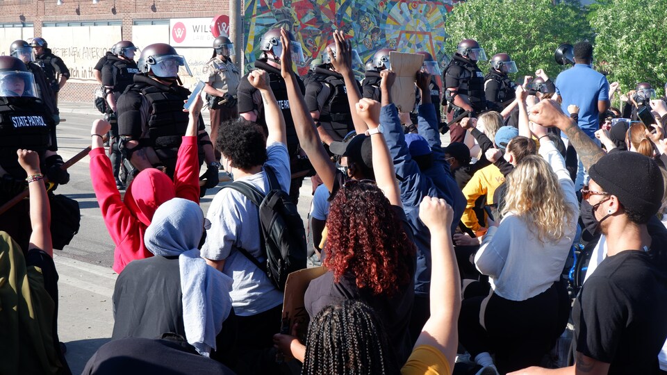 De jeunes manifestants noirs et blancs lèvent un bras face à un groupe de policiers antiémeutes.