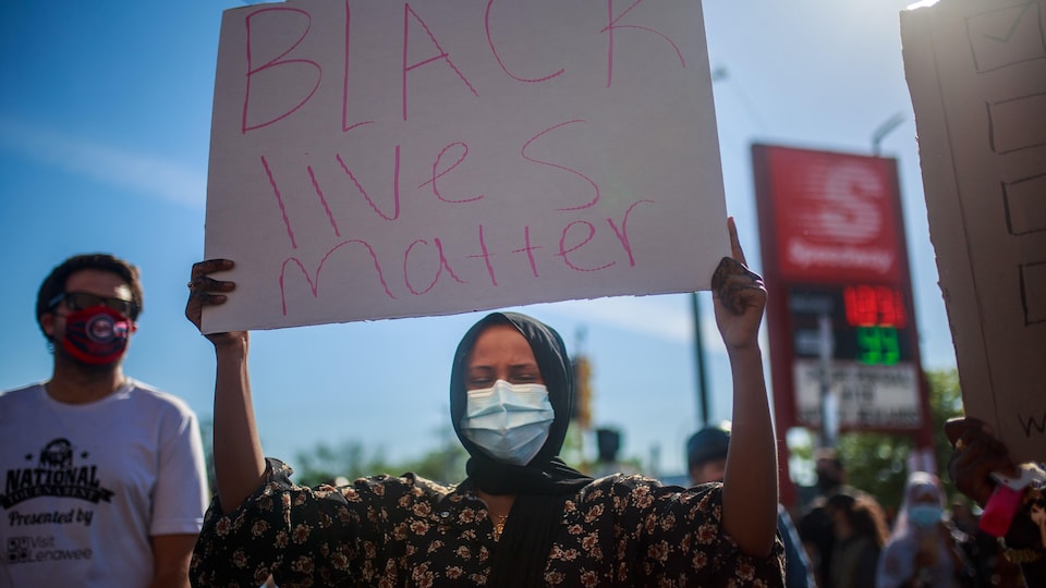 Une femme portant un masque médical brandit une pancarte sur laquelle il est écrit: «Black Lives Matter».
