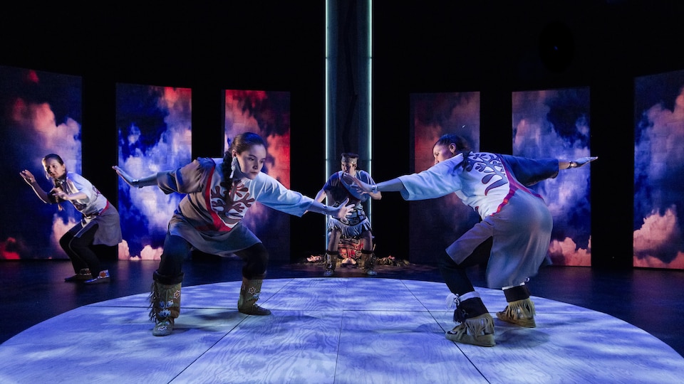 Des danseurs en habits traditionnels autochtones sont sur une scène illuminée de bleu et de pourpre.