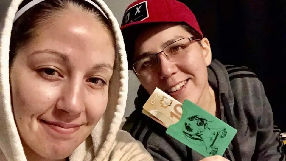 Deux personnes prennent un selfie. L'une exhibe une enveloppe verte avec une illustration de chien dessus. Un billet de 100 $ dépasse de l'enveloppe.