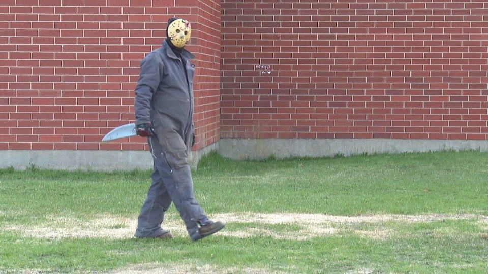 Un homme déguisé en Jason du film «Friday the 13th» marche le long d'un bâtiment. Il a un masque de hockey, une salopette grise et un faux couteau.