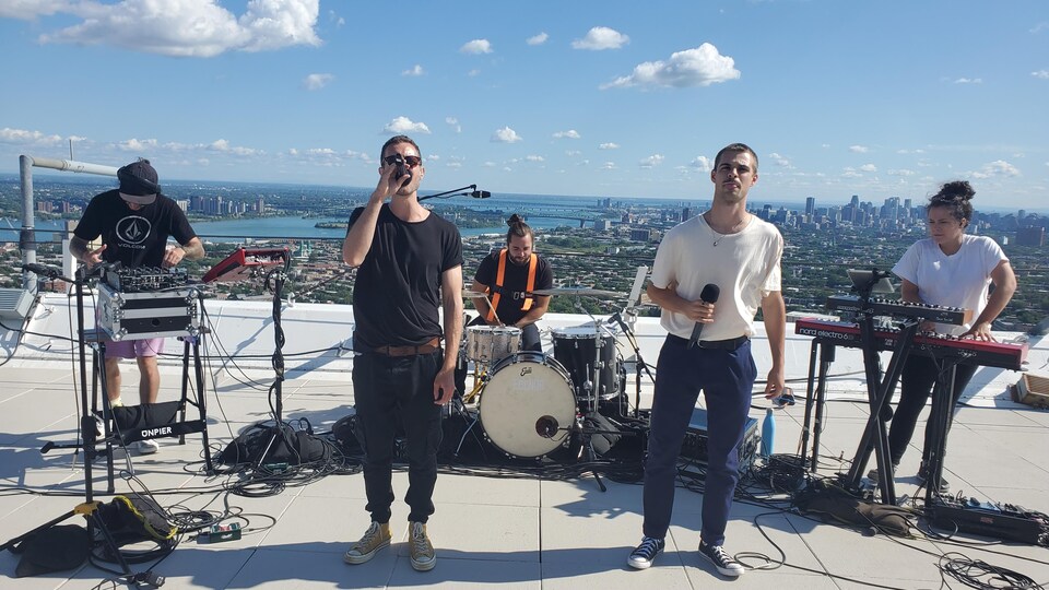 Le chanteur tient un micro et est entouré de musciens et musicienne.