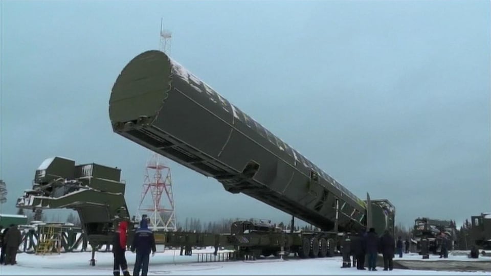 Le missile, dans un immense contenant de métal vert, est transporté à l'extérieur sur un camion remorque sous le regard de personnes vêtues de manteaux.