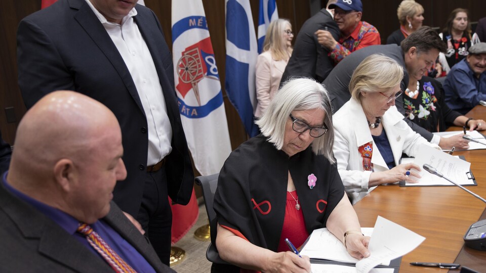 Audrey Poitras, Carolyn Bennett et d'autres personnes signent des papiers. Les drapeaux de la Nation métisse sont derrière eux.