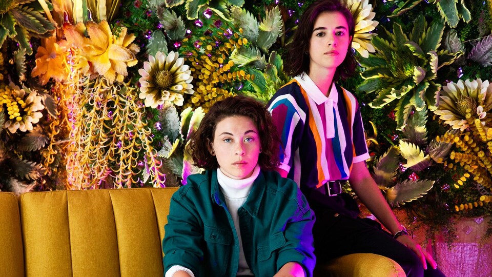 Les deux jeunes femmes sont assises côte à côte sur un sofa de couleur moutarde avec des fleurs derrière.