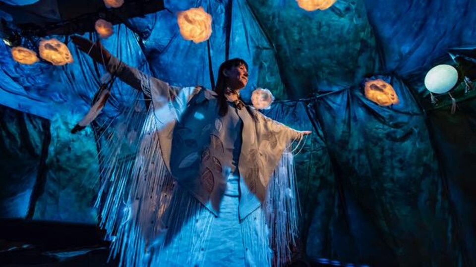 Une femme portant un costume traditionnel autochtone est debout au milieu d'une tente baignée de lumière bleutée.