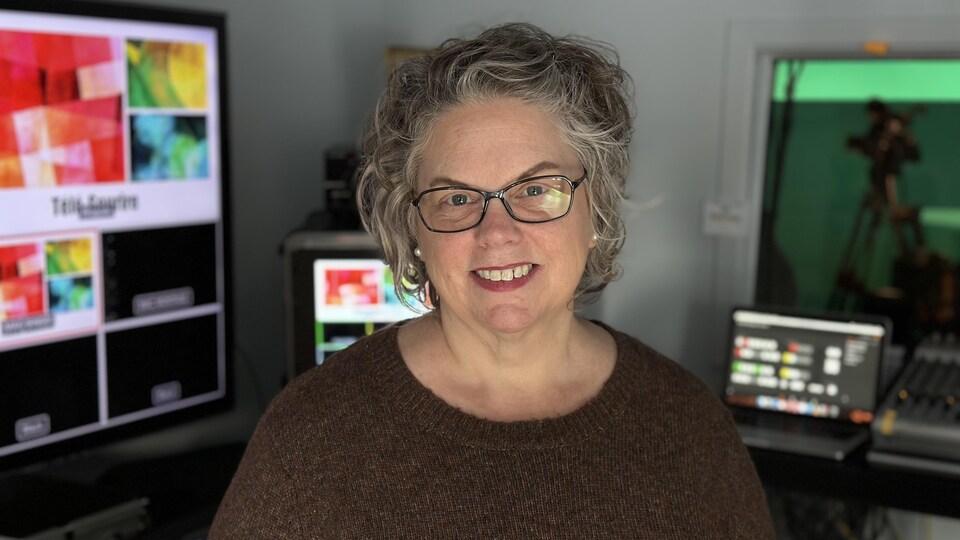 Une femme portant des lunettes souriant à la caméra dans une régie de télévision communautaire