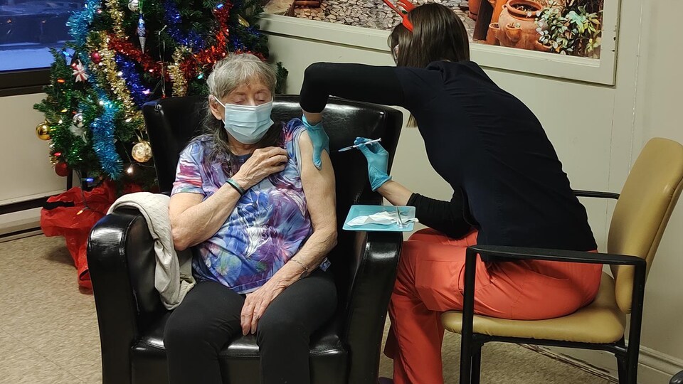 Une personne âgée, assise dans un fauteuil, reçoit un vaccin d'une professionnelle de la santé.
