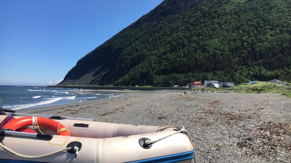 Un bateau pneumatique repose sur une plage. On voit que la plage est bordée par une grande montagne et quelques maisons. Des vagues viennent toucher la rive.