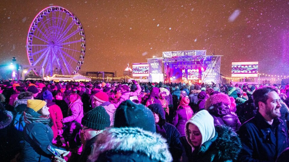 Une foule devant une scène et la roue du Vieux-Port de Montréal, à la tombée de la nuit alors qu'il neige. 