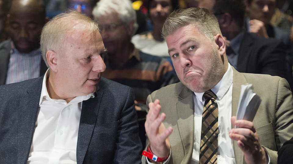 À gauche, Jean-François Lisée, la mèche tombante, et le regard attentif, regarde à droite de l'image Pascal Bérubé, cheveux coiffés vers le haut, qui fait une moue dubitative.