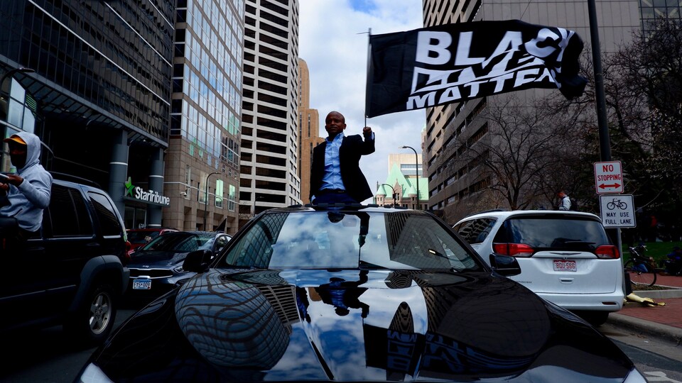 Un homme noir perché sur une voiture brandit un drapeau « Black Lives Matter ».