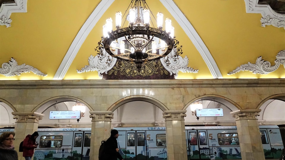 Une rame de métro arrive dans la station Komsomolskaya, la plus photogénique de Moscou, avec ses colonnes de marbre, ses fresques au plafond et ses moulures blanches sur fond jaune.