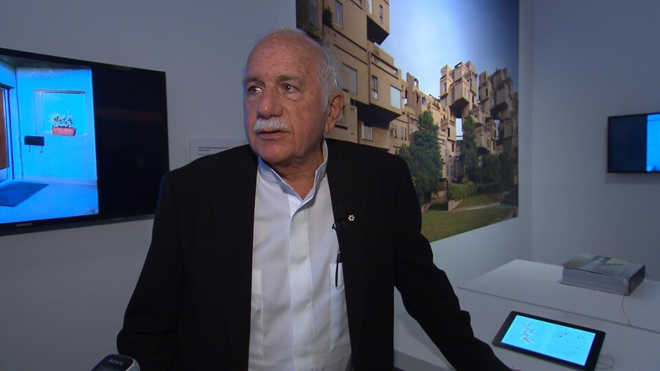 L'architecte Moshe Safdie, dans le décor d'une exposition réalisée sur son oeuvre Habitat 67, dont on voit une photo derrière lui.