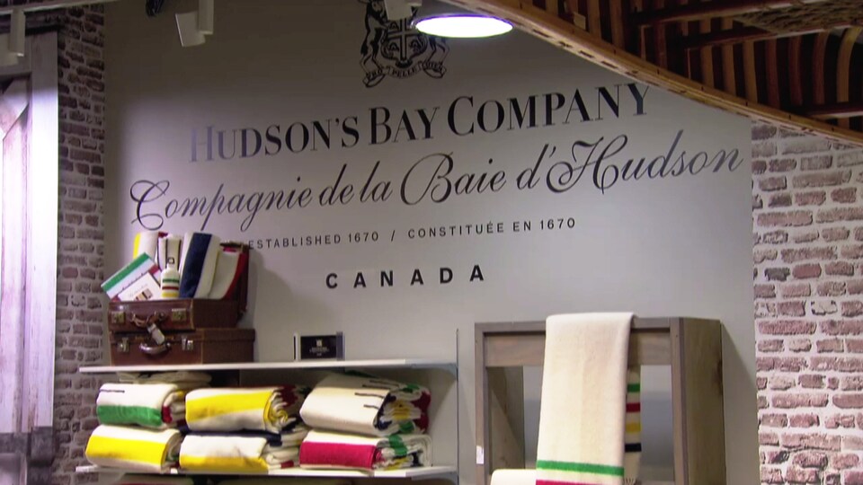 Sur un mur beige, il est écrit La Compagnie de la Baie d'Hudson. En avant, des étals offrent des produits aux couleurs de l'entreprise. 