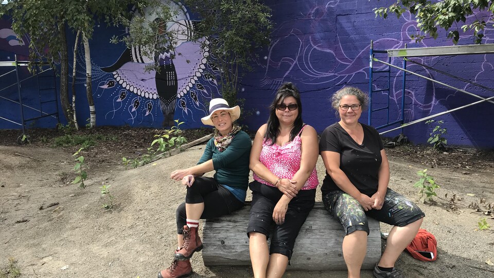 Les trois artistes sont assises sur une roche devant leur murale.