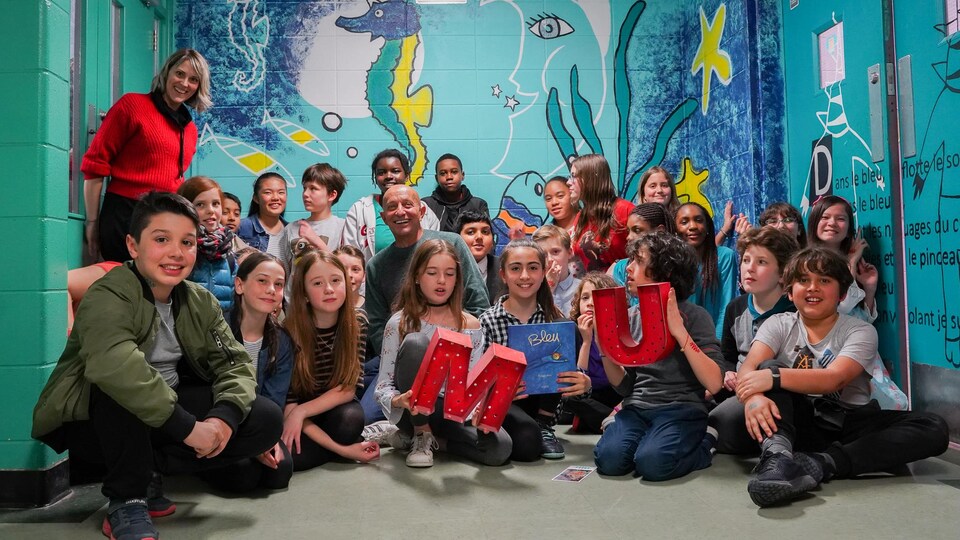 Photo en couleur d'un groupe d'enfants de 5e année du primaire devant une murale peinte sur un mur de leur école. En arrière plan, on voit sur la murale un hippocampe licorne et un croissant de lune.
