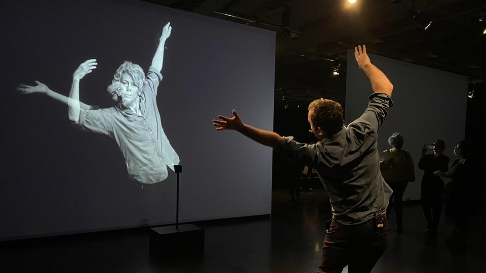 Un homme de dos agite les bras dans les airs et la danseuse sur l'écran face à lui fait sensiblement les mêmes gestes. 