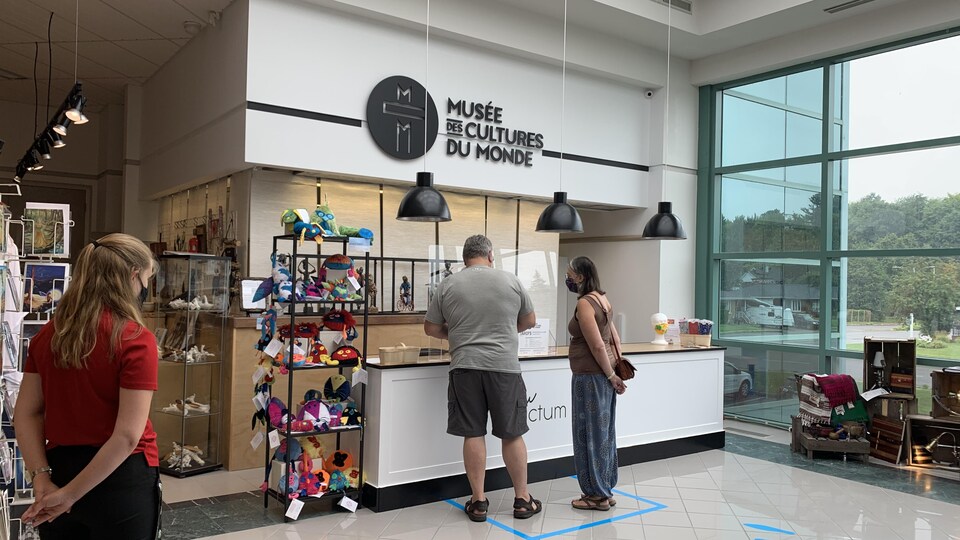 Des visiteurs sont au kiosque d'informations pour acheter un billet d'entrée. Une employée les attend à l'autre porte, les mains dans le dos. Tous portent un masque.