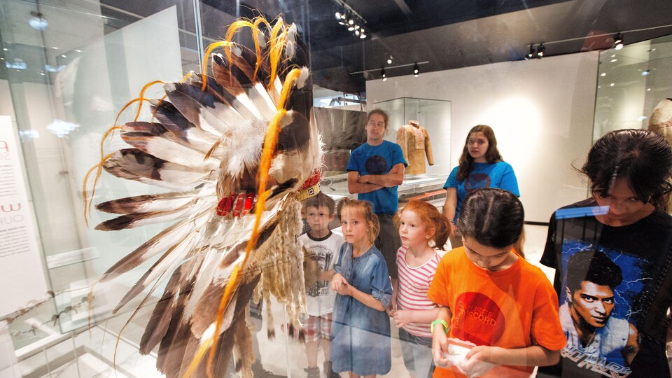 Des enfants observent une coiffe autochtone traditionnelle derrière la vitre d'un musée.