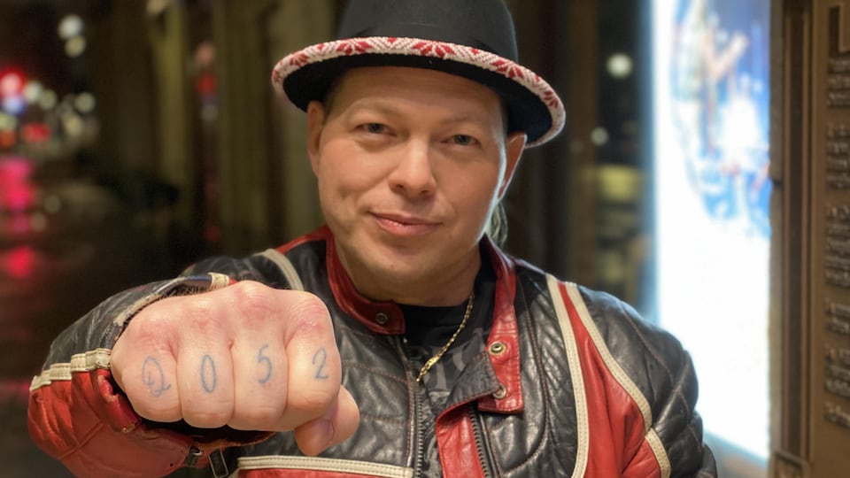 Quentin Condo avec un chapeau dont la bordure est perlée, le point tendu et un tatouage sur ses doigts avec l'inscription Q052, son nom d'artiste. 
