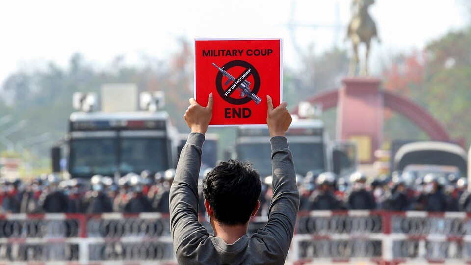 Un homme photographié de dos tient une pancarte demandant la fin du coup d'État devant des policiers protégés par des barricades.