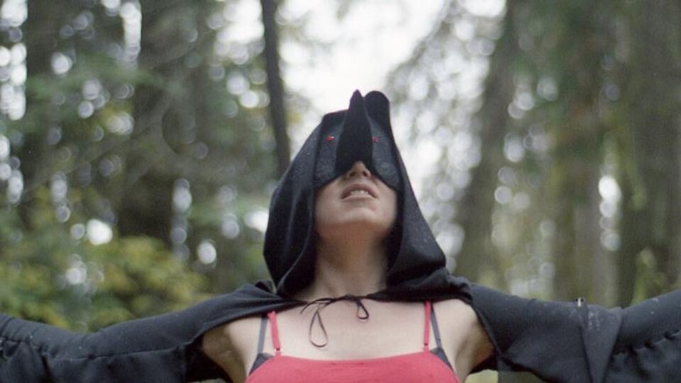 Myriam Parent ouvre les bras au beau milieu de la forêt. Elle porte un masque noir avec un long nez et des yeux rouges, et elle porte une robe rouge.