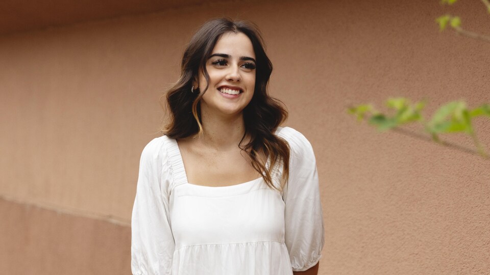 Nadia Sylvain porte une robe blance et elle sourit devant un mur dans une ruelle en vue du spectacle Polyfonik 2023.