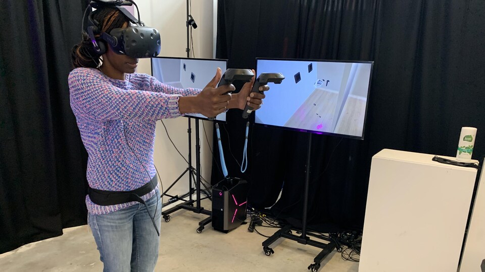Une femme avec des lunettes de réalité virtuelle et des manettes dans une salle avec des écrans.