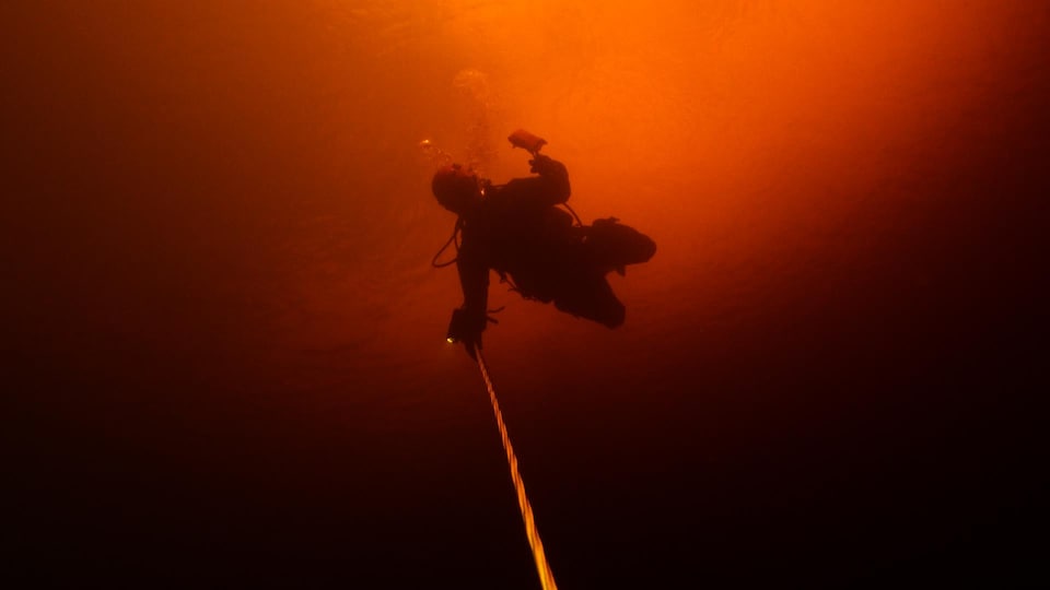 Une plongeuse en contre plongée dans des eaux sombres tient un câble. 