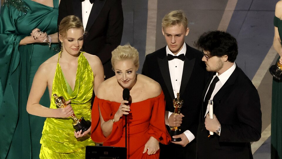 Une femme en robe de gala rouge parle au micro sur la scène des Oscars, accompagnée d'une femme et de deux hommes en smoking. 