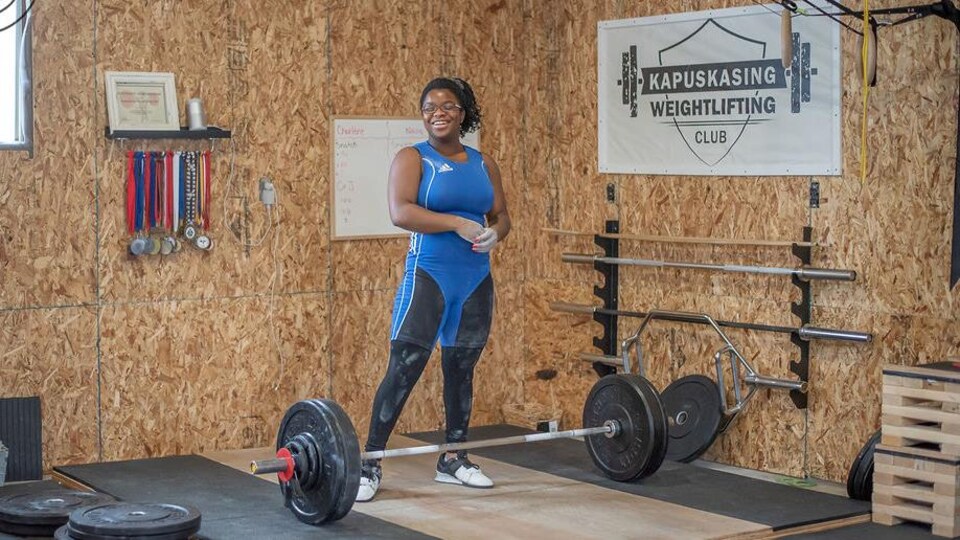 Naza Grant sourit, un haltère à ses pieds, dans un gymnase.