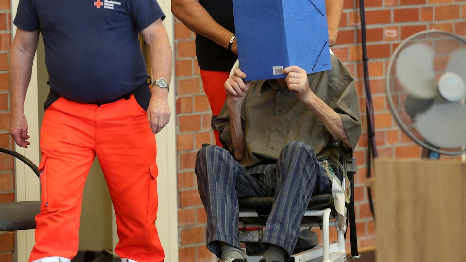 L'homme, en fauteuil roulant, se cache le visage avec une pochette. 