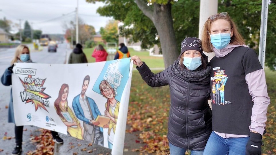 Trois femmes portant des masques tiennent une banderole où on peut lire : « Il faut que ça change maintenant! ».