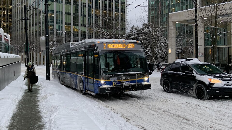 Un autobus circule sur une rue enneigée du centre-ville de Vancouver et un piéton marche sur un trottoir.