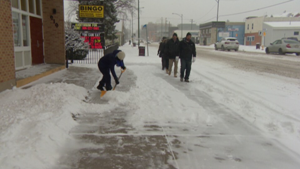 Un homme utilise une pelle pour soulever la neige et déblayer le trottoir, alors que des gens marchent près de lui. 