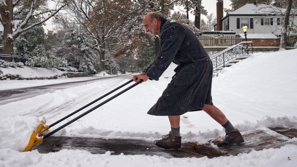 Un homme en peignoir pousse la neige dans son entrée avec une pelle.