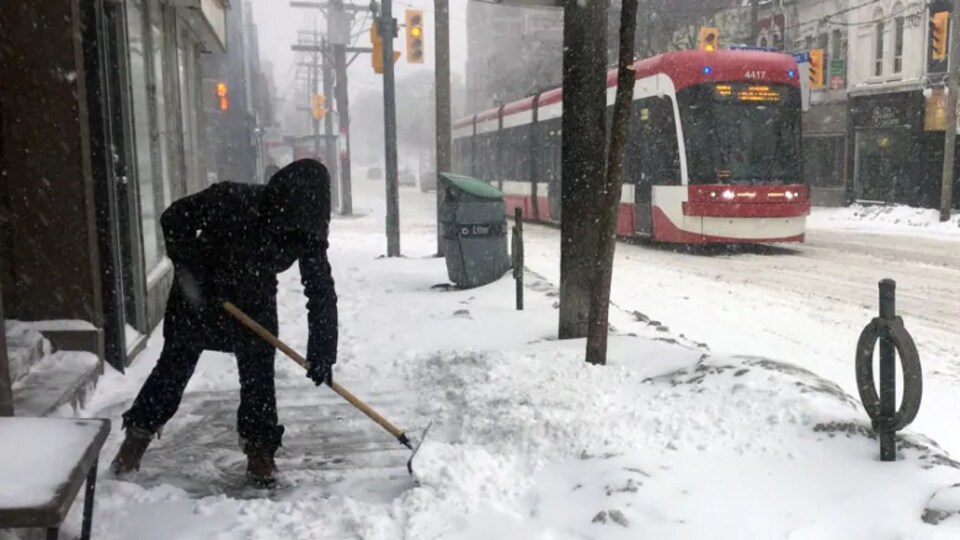 Une personne déneige le trottoir à Toronto alors qu'un tramway passe dans la rue.