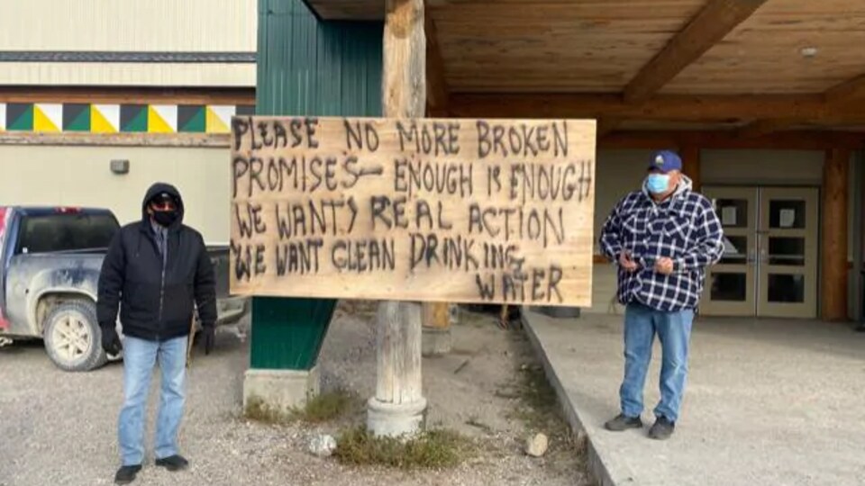 Deux hommes de chaque côté d'une pancarte sur laquelle est écrit « S'il vous plaît, plus de promesses brisées, assez c'est assez. »