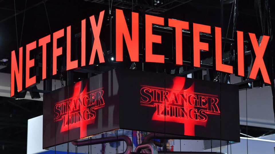 Deux ensembles de lettres géantes composant le mot Netflix sur une structure suspendue, avec en dessous deux écrans projetant le logo de Stranger Things, saison 4. 