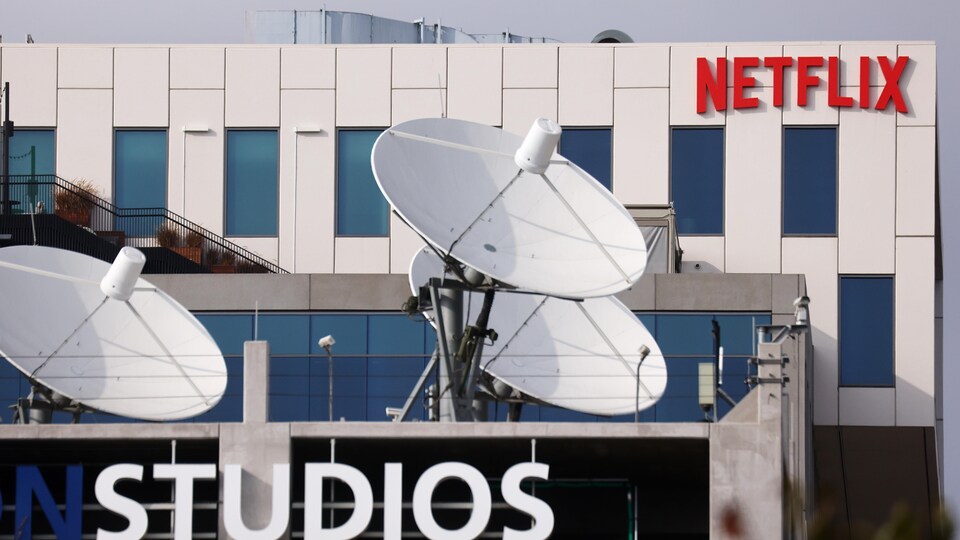 Le logo de Netflix est visible au sommet de la façade d'un grand édifice blanc. Devant l'édifice, on aperçoit d'imposantes antennes paraboliques.
