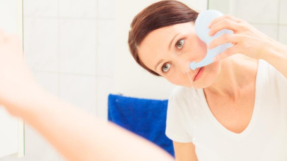Une femme utilise un pot de douche nasale en se regardant dans le miroir.