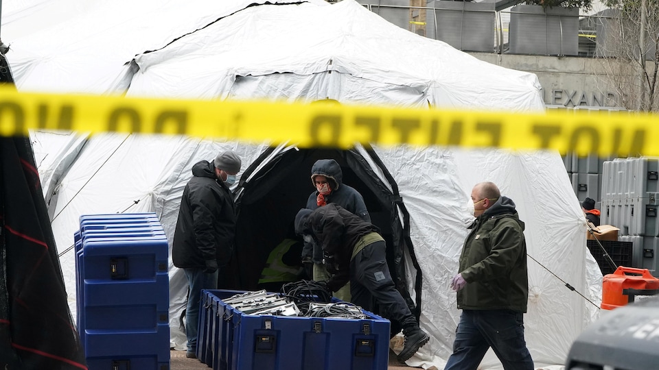 Des hommes portant de masques transportent du matériel dans une tente blanche. Un ruban limite l'accès au site.